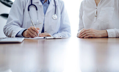 Doctor and patient sitting near each other at the wooden desk in clinic. Female physician's filling up a records form. Medicine concept - 625988282