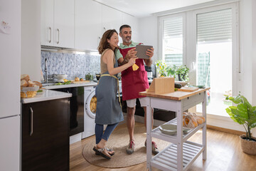 wide shot of an adult caucasian couple in the middle of the kitchen. They are standing in their aprons smiling and looking at a recipe on their tablet
