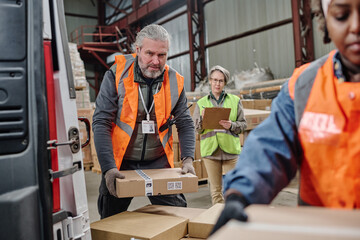 Mature worker looking at camera while loading boxes in truck together with his colleagues