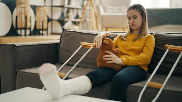 Teen girl with plaster on feet sit on bed with help crutches. Young woman sit in bed and holding teddy bear in arms, speak complains of pain and strokes him. Sadness and loneliness of sick children.