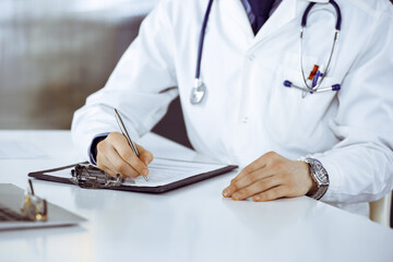 Unknown male doctor sitting and working with clipboard of medication history record in clinic at his working place, close-up. Young physician at work. Perfect medical service, medicine concept