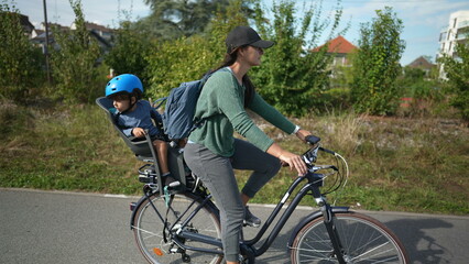 Fototapeta na wymiar Mother riding bicycle with child in back seat outside in urban green path