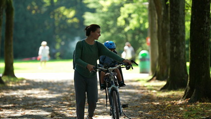 Mother carrying bicycle outside at park with child on back seat bike