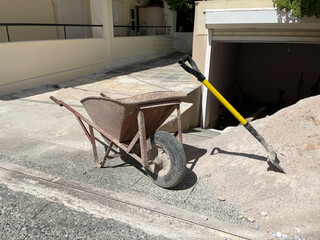An empty old rusty hand wheelbarrow and shovel next to the pile of sand. A city street, contraction...