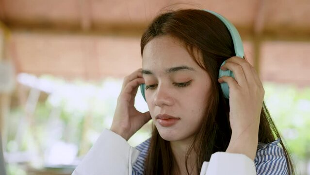 Close-up Asian teenage woman face, is wearing blue headphones listen music when wants relax, from typing work on laptop computer, sit listen chill music in restaurant on holiday that wants relax.