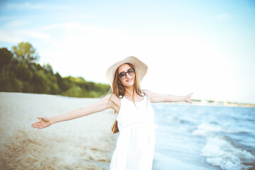 Fototapeta na wymiar Happy smiling woman in free happiness bliss on ocean beach standing with a hat, sunglasses, and open hands. Portrait of a multicultural female model in white summer dress enjoying nature during travel
