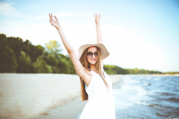 Fototapeta na wymiar Happy smiling woman in free happiness bliss on ocean beach standing with a hat, sunglasses, and rasing hands. Portrait of a multicultural female model in white summer dress enjoying nature during