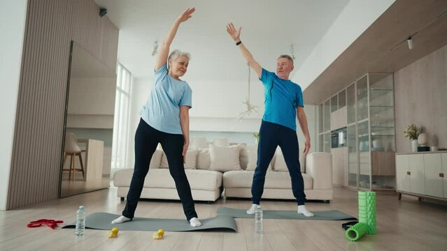 Elderly couple man and woman exercising together at home in living room. Senior man and woman doing fitness together. Aged people leaning to sides standing on mats. Sport, training, workout concept.