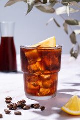 Cold brew coffee in a glass with ice and lemon on a light marble background with coffee beans and bottle close up. Concept summer craft refreshing homemade drink.
