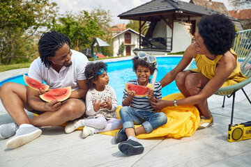 Beautiful and joyful african american family of four sitting by the pool and eating watermelon together.