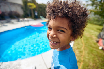 Portrait shot of lovely african american boy standing by the outdoor in ground pool in the backyard, smiling and feeling happy. - 626013669