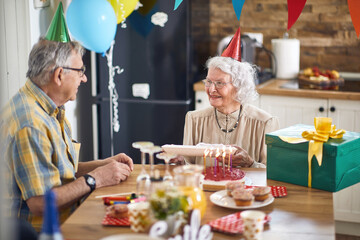 Beautiful joyful senior couple with birthday hats sitting at kitchen table together celebrating senior womans birthday.