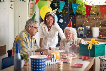 Grandma Blows Out Candles Surrounded by Grandpa and Caregivers Amidst Falling Confetti