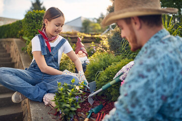 Beautiful young girl using garden rake sitting by the garden outdoors planting flowers with her father on a sunny summer day.