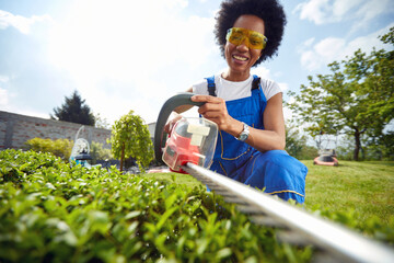 Joyful young woman in working clothes using electric bush trimmer on a sunny summer day outdoors in...