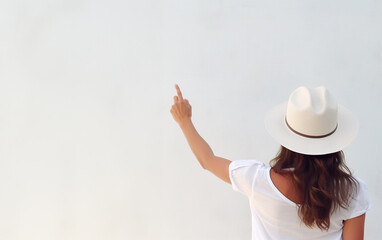 A young woman in summer clothes stands in front to the white wall, his hand raised and pointing a point on the wall