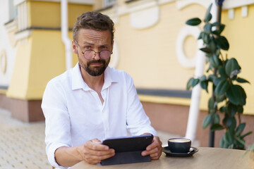 Funny man sits at table, engrossed in reading news on tablet PC. Face displays surprised...