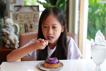 Asian girl child sitting by table and eating Blueberry Cheesecake cake in Cafe. Kid eats tasty food.