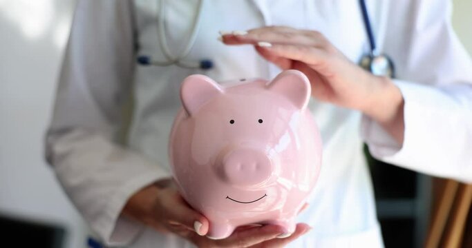 The doctor in a white uniform is stroking a piggy bank, a closeup. The cost of counseling with a medical specialist