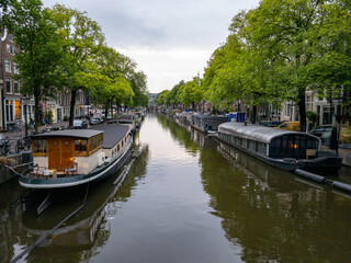 Fototapeta na wymiar Houseboats line a tree-lined canal with calm water and reflections in the city of Amsterdam