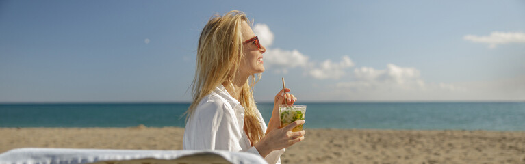 Side view of happy woman enjoying mohito cocktail and smiling away on empty beach