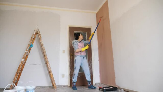 wall painting. a woman, in work clothes, paints a white wall indoors with pink paint using a roller on a long holder.