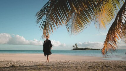 Woman walking barefoot white sand beach at sunset. Tourist girl in black dress enjoy tropical island landscape, blue ocean water and coconut palms. Travel, tourism, holiday. Back view, slow motion