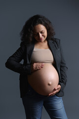 Elegant multi-ethnic pregnant woman smiles while gently caressing her big belly in pregnancy 36 week, feeling baby kicks