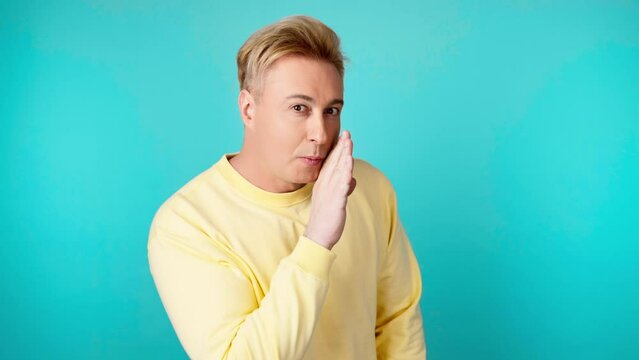 Secret, mystery and man keeps index finger on lips, makes silence gesture. Positive expression, confident smiling guy in yellow sweatshirt looks around in studio isolated on blue background.