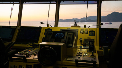 Main console of navigational bridge of modern ultra large container vessel with different data screens and navigational wheel during sunset 