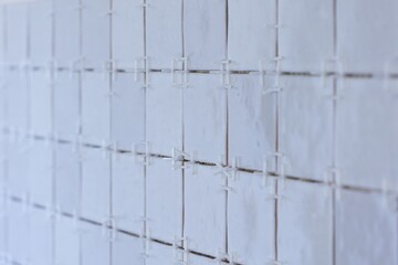 A portrait of a tile wall under construction. The wall is being tiled with small ceramic rectangular tiles. There are still white plastic joint spacers in between the junctures of the tiles.