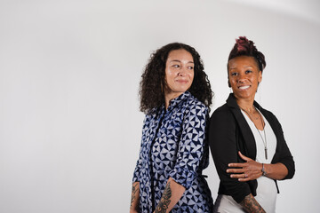 Two women stand shoulder to shoulder. One of them is in a neat black blazer, and the other is in a patterned dress. One woman looks over her shoulder to look at the other, who smiles at the camera.