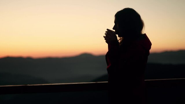 Silhouette of young woman traveler enjoying cup of coffee or tea on warm sunset or sunrise in the mountains Girl taking a sip of coffee or tea with incredible view on the background