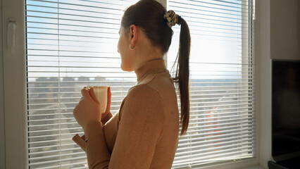 Beautiful woman drinking coffee and opening window blinds. People in morning, beautiful cityscape,...