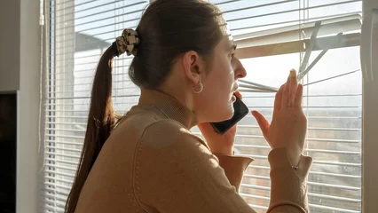 Foto op Plexiglas Young worried woman talking by phone and looking through window blinds. Crime witness, spying through window, peeking on street. © Кирилл Рыжов