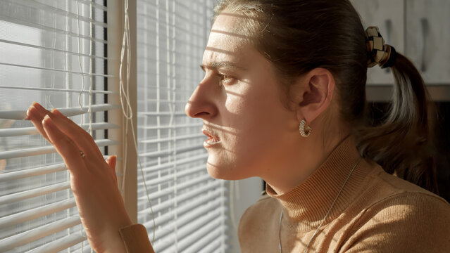 Portrait of shocked scared woman looking outdoors through window. Crime witness, spying through window, peeking on street.