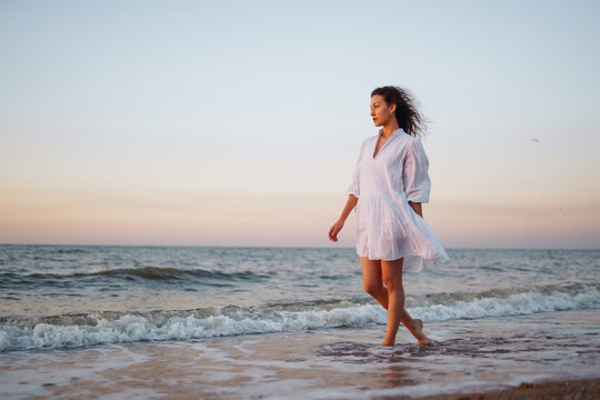 Slender young woman in airy white is enjoying the sunset light on the seashore. Summer time. Travel, weekend, sunny, fun, positive mood. Active lifestyle.