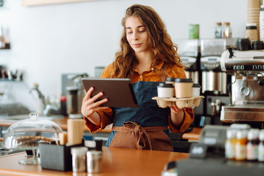 Handsome young manager of a coffee shop with curly hair and a tablet in his hands gives takeaway coffee in a coffee shop. Takeaway food. Business concept.