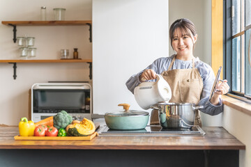 Fototapeta na wymiar キッチンで電気ケトルのお湯を鍋に入れるエプロン姿のアジア人女性 
