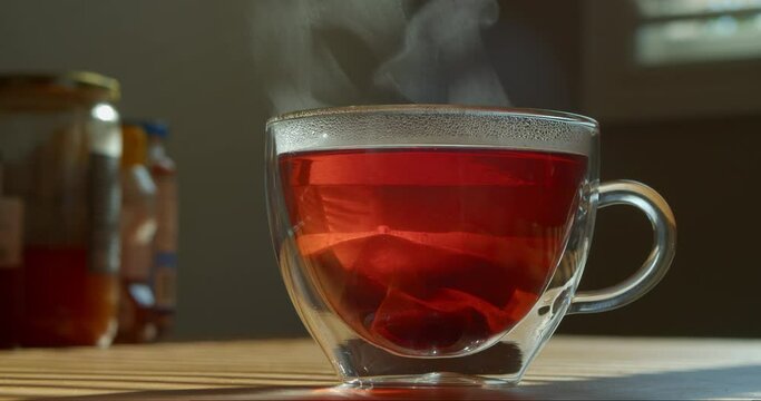 Hot tea in a transparent cup on the kitchen table by the window. Cup of freshly brewed black tea, steam coming out, warm soft light from the window