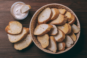 Pieces of roasted white bread in wicker basket and glass bowl of sauce on a dark background. Closeup of delicious croutons. Top view.