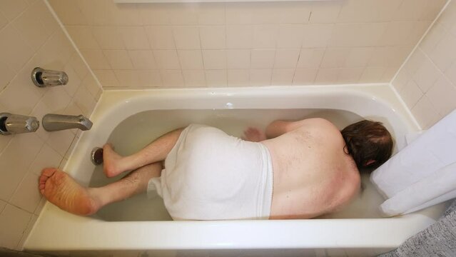 Man curled and submerged face down in bathtub full of water as if playing dead.