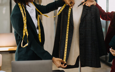 Female fashion, designer, Looking at Drawings and Sketches that are Pinned to the Wall Behind Her Desk. Studio is Sunny. Personal Computer, Colorful Fabrics, Sewing Items are Visible.