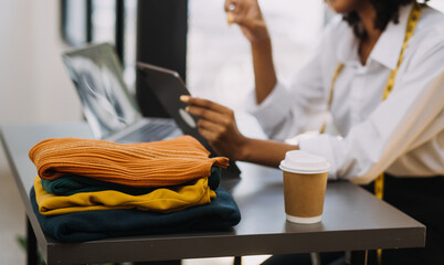 Female fashion, designer, Looking at Drawings and Sketches that are Pinned to the Wall Behind Her Desk. Studio is Sunny. Personal Computer, Colorful Fabrics, Sewing Items are Visible.