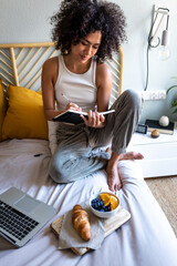 Multiracial latina woman writing daily affirmations on journal while eating breakfast in bed next...
