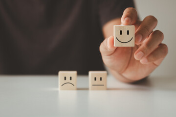 Customer satisfaction concept. Customer's hand lifts smiling face on blurred sad face icon on wooden cube block, Service rating, customer service best excellent business rating experience survey.