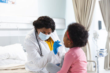 African american female doctor wearing face mask examining girl patient at hospital