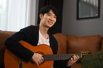 Obraz premium Pleasant asian man playing acoustic guitar on couch, spending leisure time in cozy living room