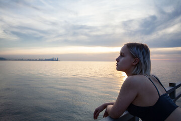 Fototapeta na wymiar Blonde woman in tank top standing on pier on the background of city silhouette, cloudy dusk sky and sea