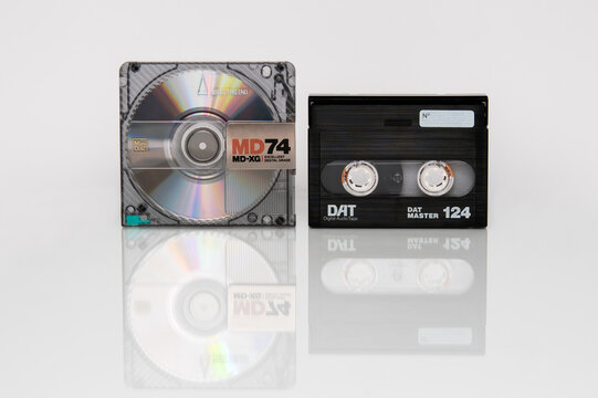 A minidisc shimmering with iridescent reflections and a DAT cassette stand side by side vertically isolated on a white background with reflection